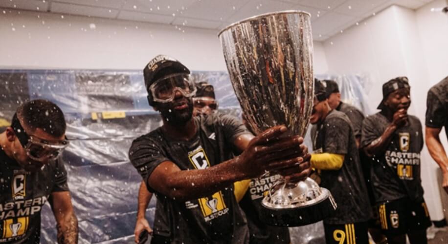 Athlete wearing goggles holding a large silver trophy, teammates celebrating around him.