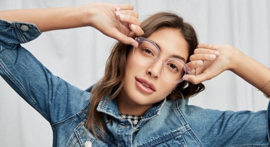 Woman wearing clear acrylic glasses and a denim jacket, adjusting the glasses with her hands.
