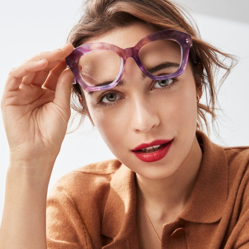 Woman wearing oversized lavender transparent glasses with a brown button-up shirt.