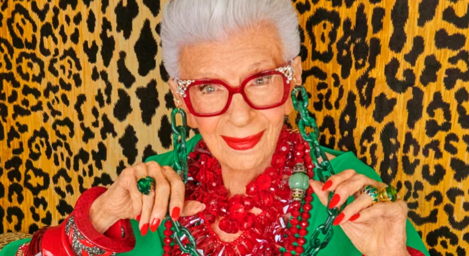 Woman wearing large red glasses, multiple red and green statement necklaces, and a green top against a leopard-print background.