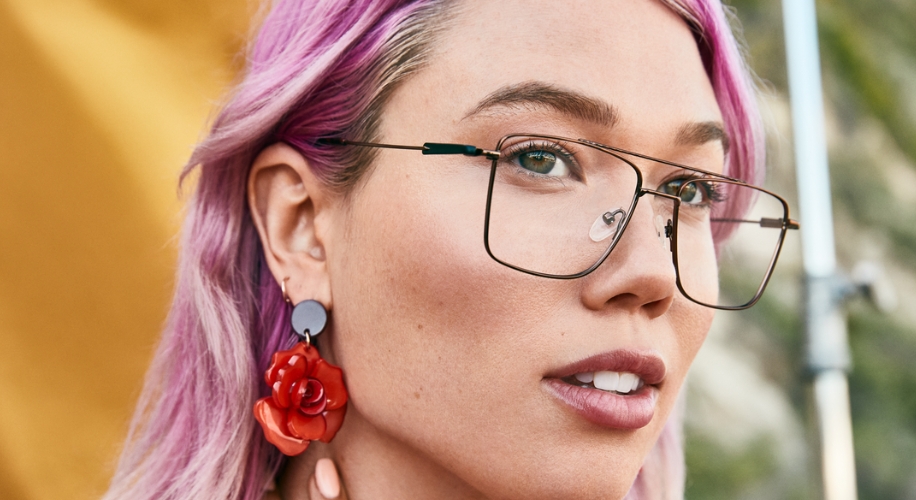 Woman with purple hair wearing large, square-framed glasses and red floral earrings.