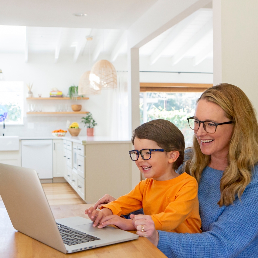 Smiling woman and a child wearing glasses using a laptop at a table.