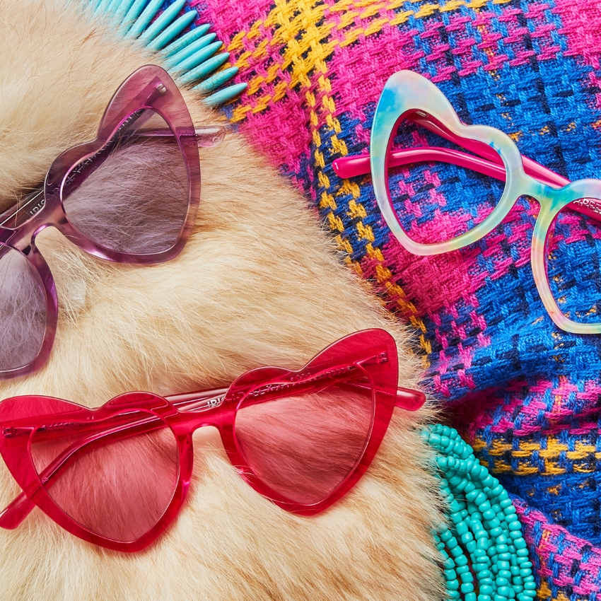 Three pairs of heart-shaped sunglasses in pink, purple, and multicolored frames arranged on fabric surfaces.