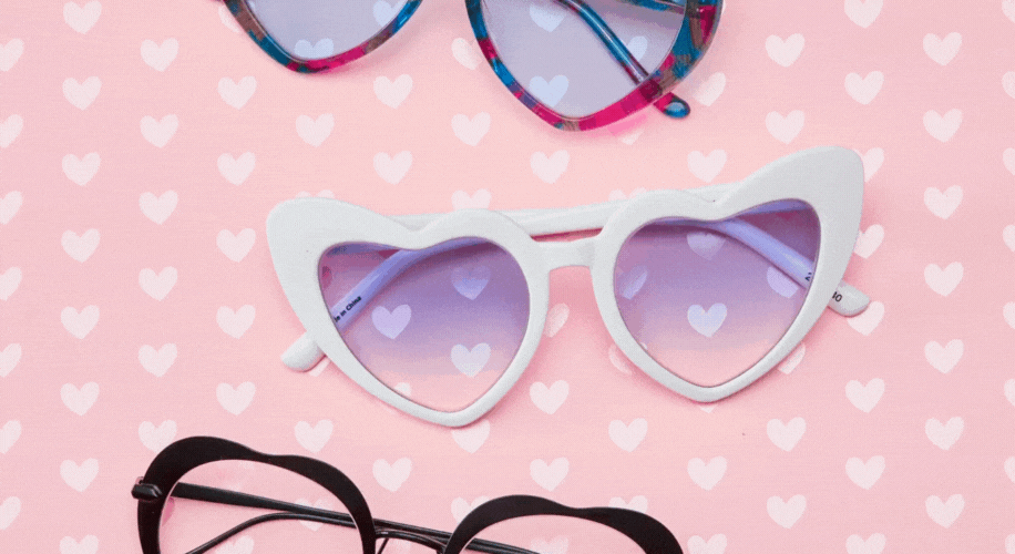 Three pairs of heart-shaped sunglasses with different colored frames on a pink heart-patterned background.