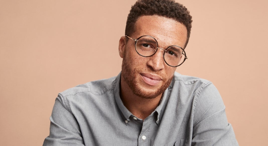 Man wearing round eyeglasses and a grey button-up shirt against a plain background.