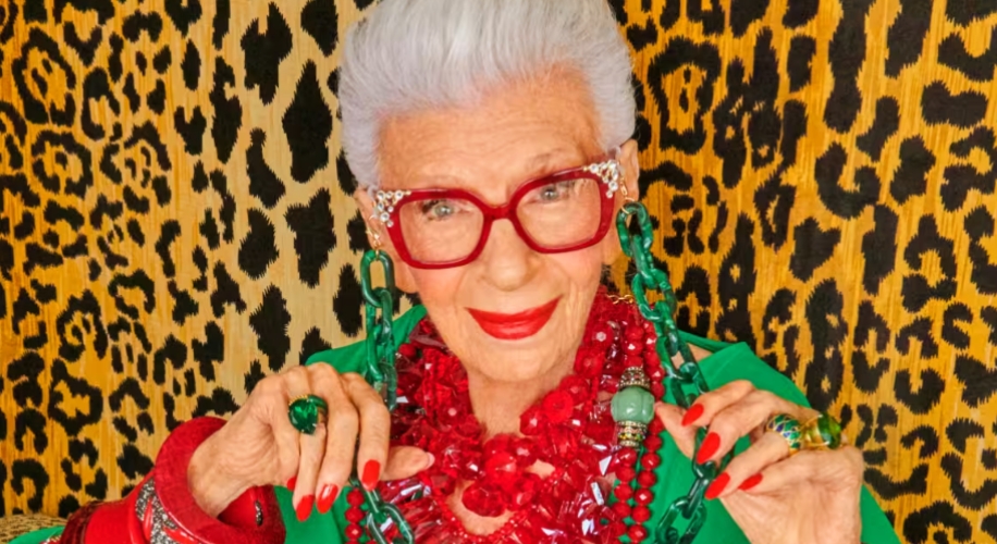 Elderly woman wearing red glasses, red necklace, green earrings, red and green rings, and red nail polish.