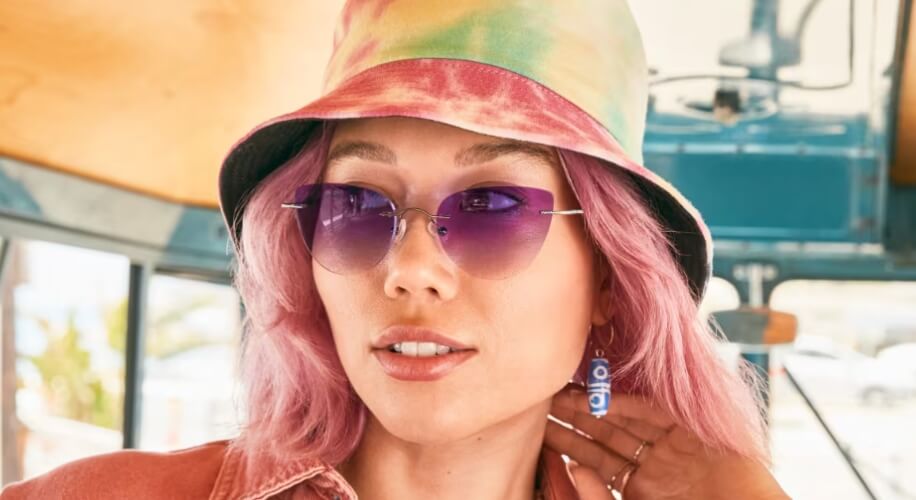 Woman wearing a tie-dye bucket hat, purple-tinted sunglasses, and colorful earrings with long pink hair.