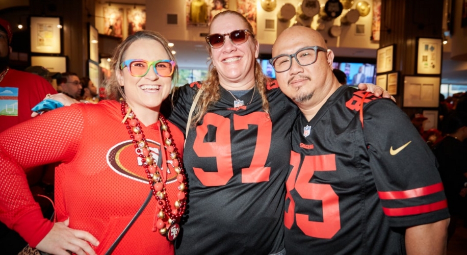 Three people in 49ers sports jerseys smiling, with the numbers 97 and 85 visible and one wearing a red top.