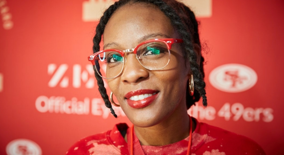 Person with twisted hair wearing red and clear-rimmed glasses, smiling with bright red lipstick.