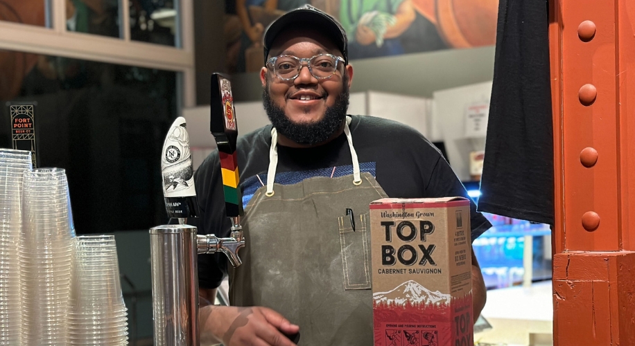 Person standing behind a counter next to a box labeled &quot;Top Box Cabernet Sauvignon.&quot;