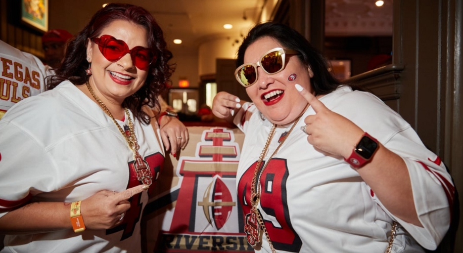 Two women wearing white jerseys with number 49, heart-shaped sunglasses, and NFL-related accessories.