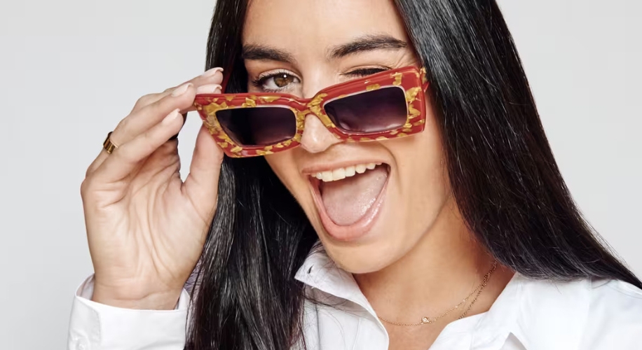 Woman with long black hair winking and smiling, wearing red and gold patterned sunglasses.