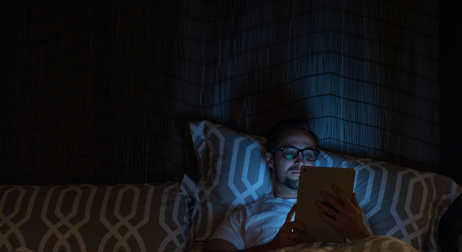 Man using a tablet in bed with patterned pillows and dim lighting.