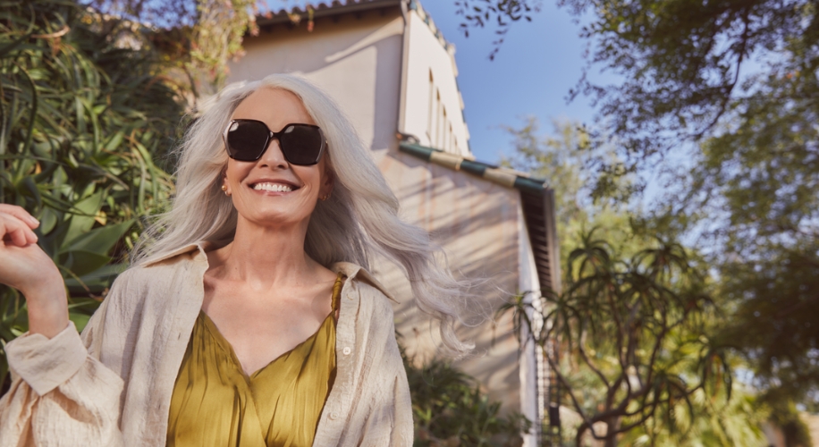 Woman wearing large black sunglasses and a light yellow blouse, smiling outdoors on a sunny day.