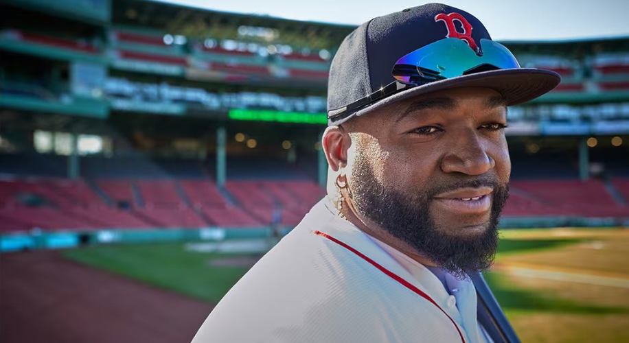 Smiling man with a beard wearing a Red Sox cap and reflective sunglasses, holding a baseball bat.