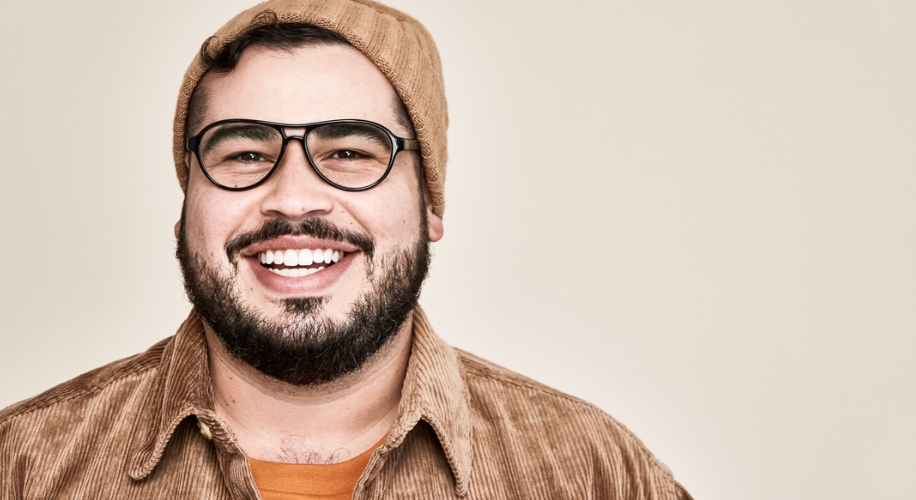 Man wearing black-rimmed glasses, a tan beanie, and a brown corduroy shirt smiling.