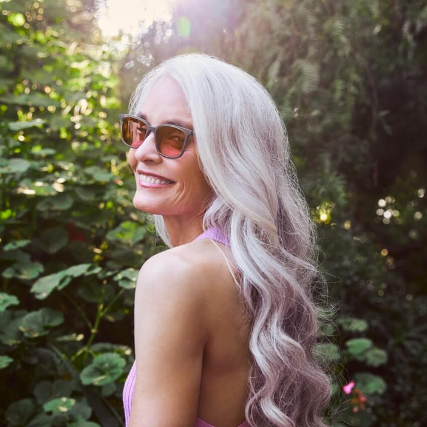 Woman with long, wavy gray hair wearing sunglasses and a pink top smiles while looking over her shoulder.