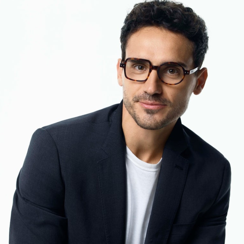 Man wearing rectangular tortoise-shell glasses and a dark blazer over a white shirt.