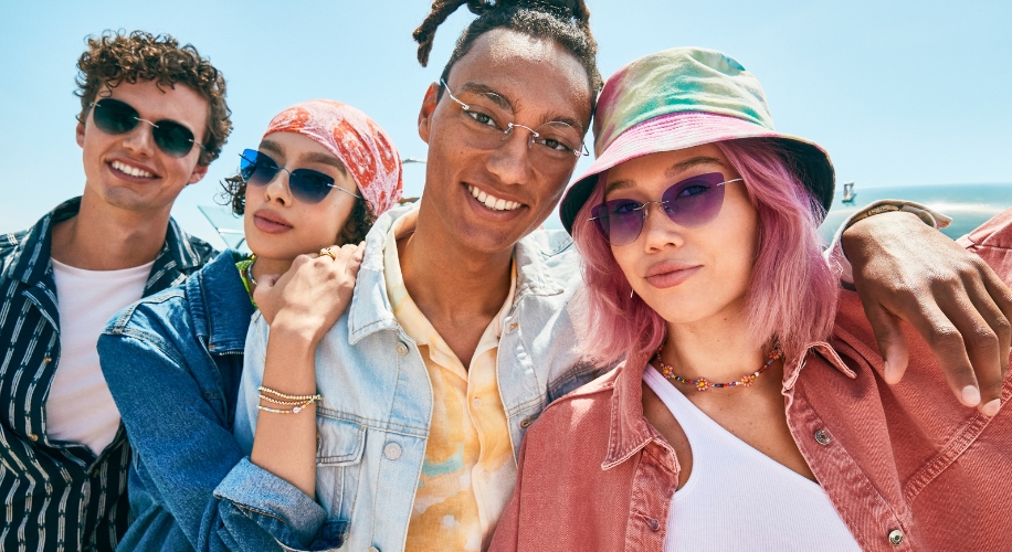 Group of four friends wearing sunglasses and casual clothing, smiling on a sunny day.