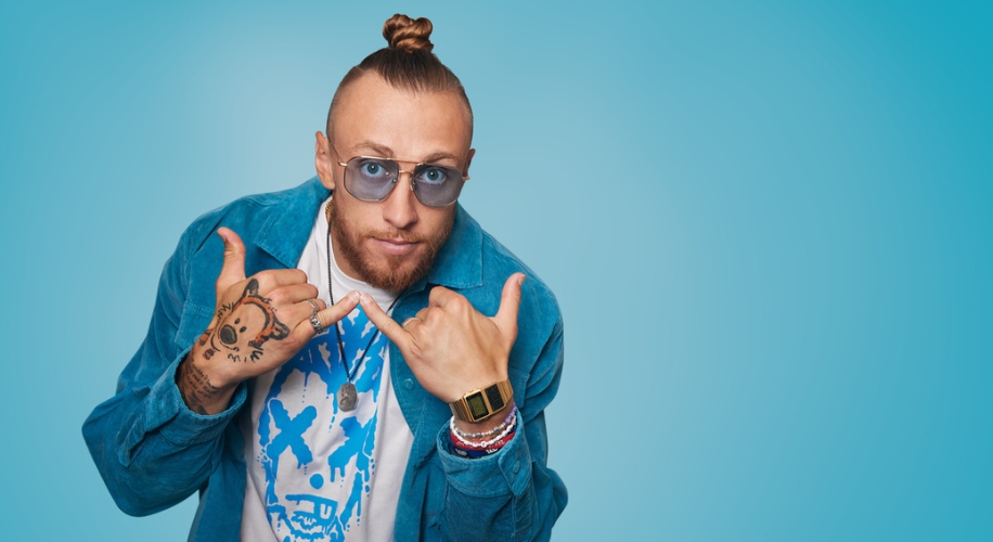 Man with top knot wearing blue glasses, a blue suede jacket, and a graphic t-shirt making a hand gesture.