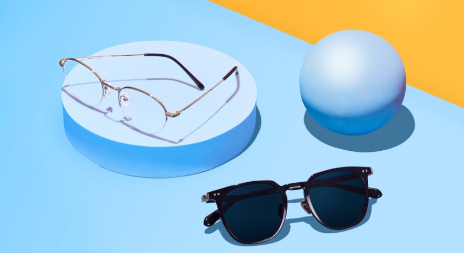 Two pairs of eyeglasses on a blue and yellow background. One is a thin-framed prescription lens, the other is black-framed sunglasses.