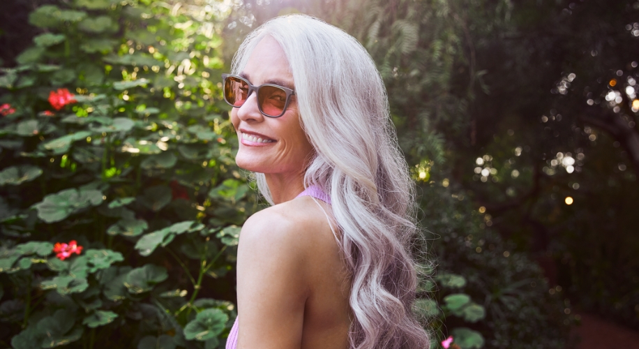 Woman with long, silver hair wearing sunglasses and a light purple top smiles outdoors in a garden.