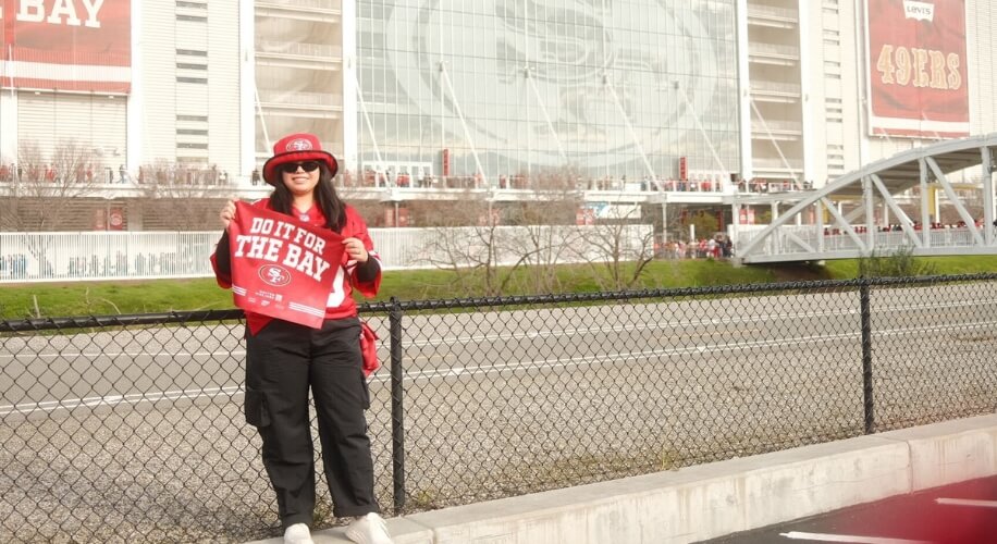 Fan holding a red &quot;DO IT FOR THE BAY&quot; banner outside a stadium, wearing a matching hat and jersey.