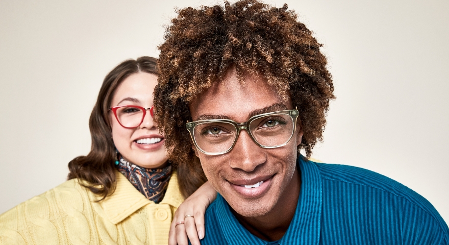 Two people smiling, one wearing red-framed glasses and the other wearing green-framed glasses.