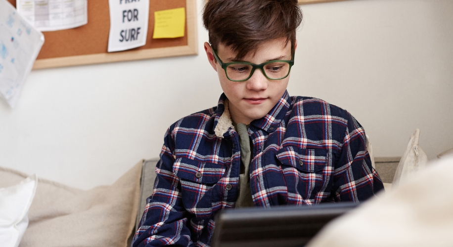 Boy in green glasses and a plaid shirt looking at a tablet screen.