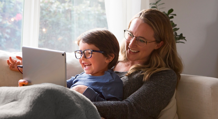 Child and adult smiling and wearing glasses, using a tablet while sitting on a cozy sofa.