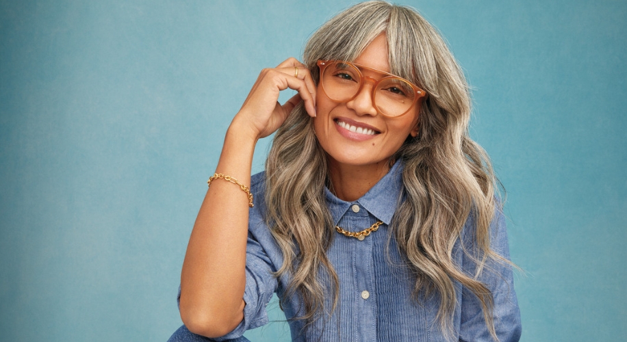 Woman modeling large orange frame glasses, wearing a blue button-up shirt and gold chain necklace.