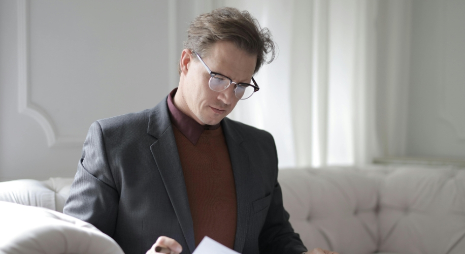 Man in a suit sitting on a white couch and reading documents attentively.