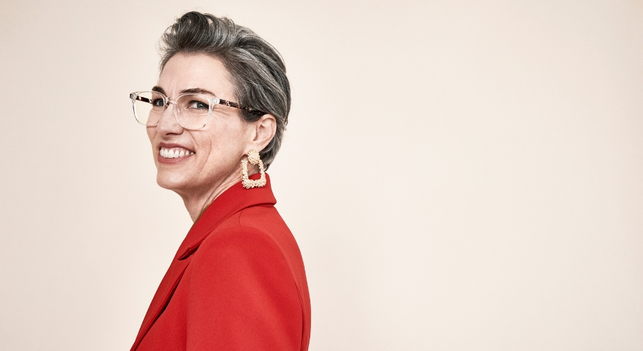 Woman in a red blazer, wearing oversized glasses and geometric gold earrings, smiling to the side.