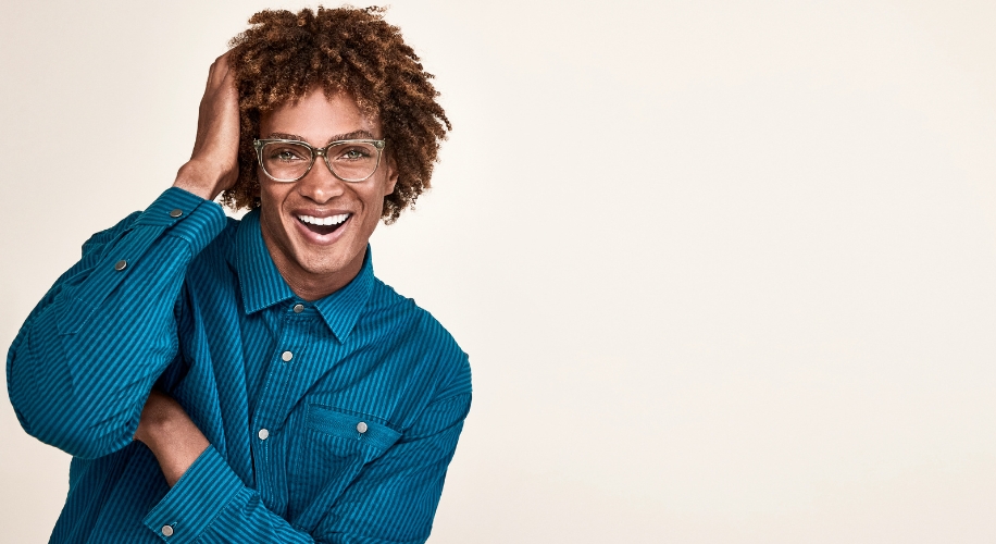 Person wearing a teal button-up shirt and glasses, smiling with one hand touching their hair.