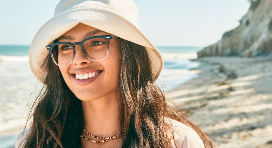 Woman wearing blue square glasses and a white bucket hat, smiling on a beach.