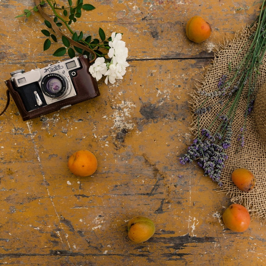 Vintage camera with leather cover, white flowers, apricots, lavender bunch, and burlap sack on rustic table.