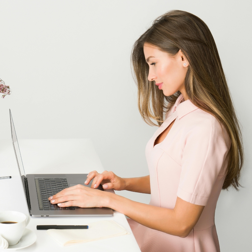 Woman typing on a laptop with a cup of coffee, pen, and napkin on the table.