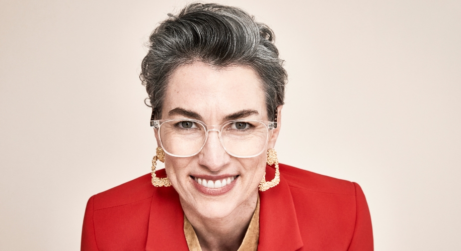 Smiling person with short gray hair wearing clear eyeglasses, gold square earrings, and a red blazer.