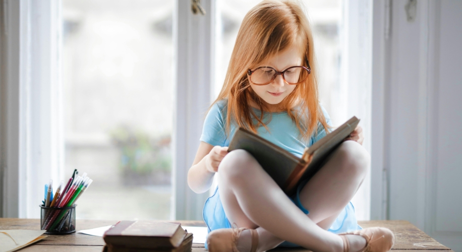 Young girl with glasses and a blue dress engrossed in reading a book, sitting cross-legged on a wooden table.