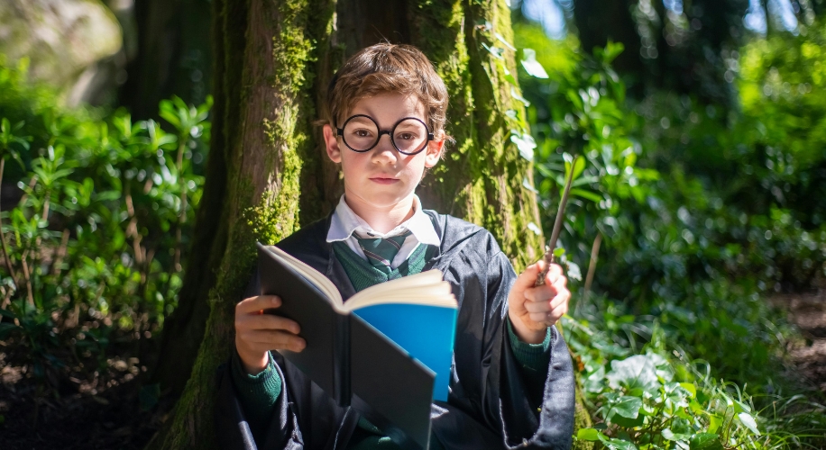 Child dressed as a young wizard reading a book and holding a wand, seated against a tree in a forest.