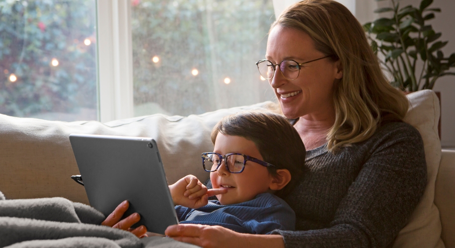 Woman and child wearing glasses, sitting on a couch, looking at a tablet together.