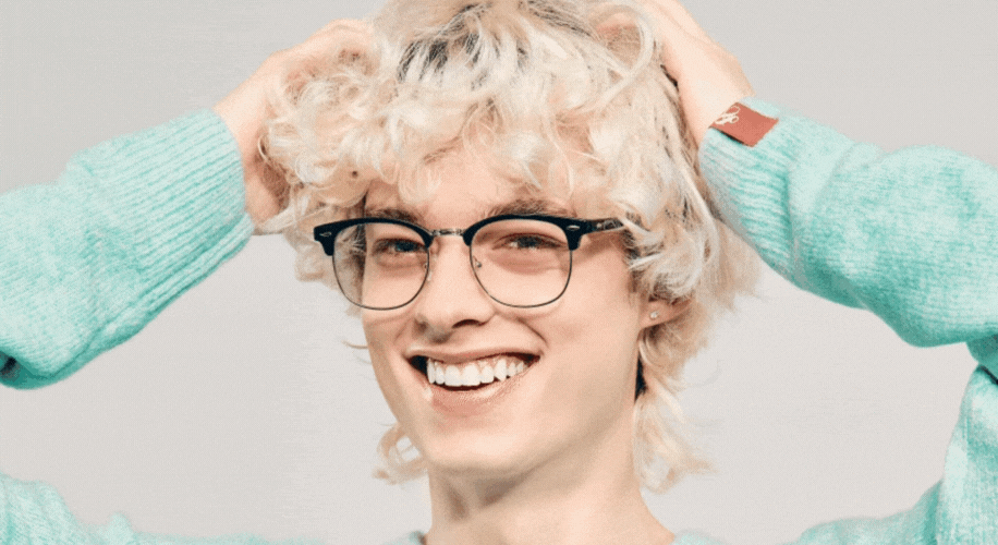 Person wearing black-rimmed eyeglasses and a light blue sweater, smiling with hands in curly blonde hair.