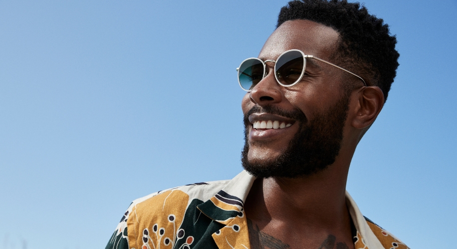 Man wearing round sunglasses and a patterned shirt with abstract designs, smiling under clear blue sky.