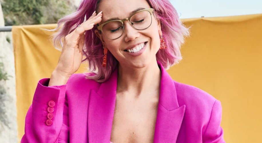 Person wearing a bright pink blazer, glasses, and red earrings, smiling with eyes closed.