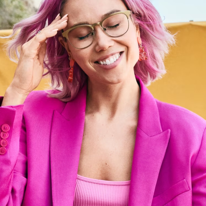Person wearing glasses, a bright pink blazer, a matching pink top, and pink earrings, smiling with closed eyes.