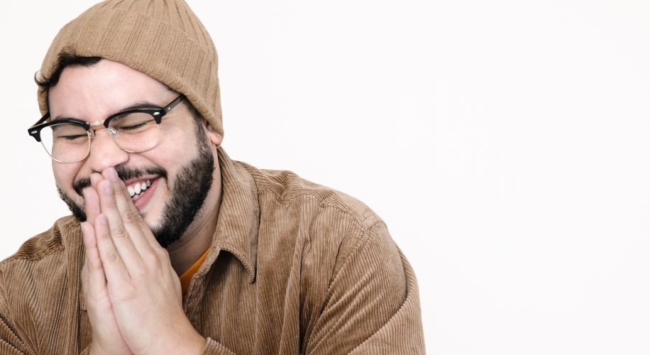 Man wearing a brown beanie, glasses, and a brown corduroy jacket, smiling with hands pressed together.