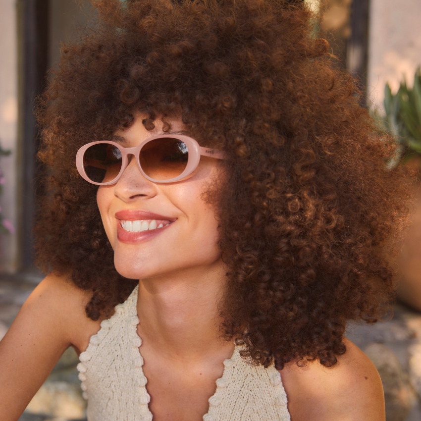 Woman wearing pink oval sunglasses with a knitted cream top, smiling.