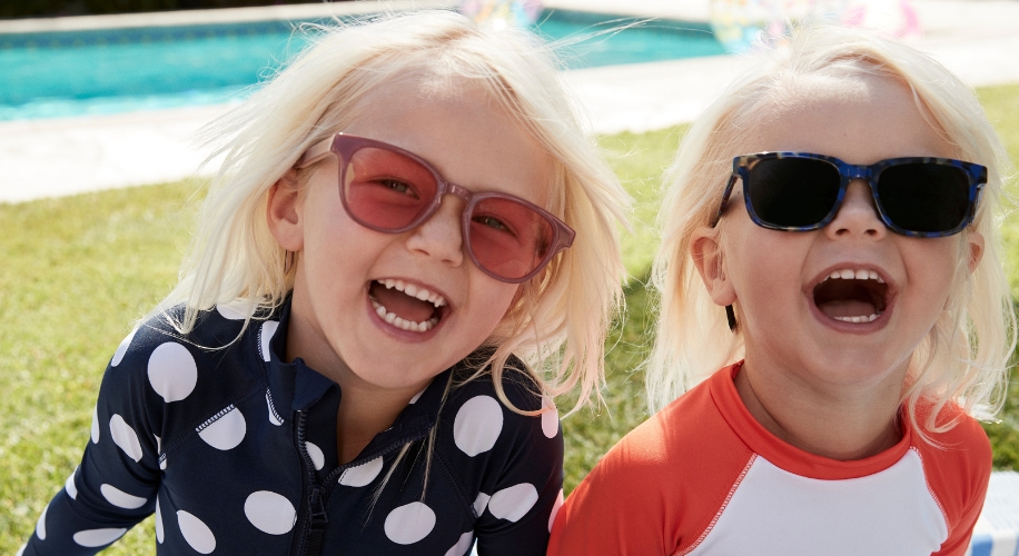 Two children wearing colorful swimsuits and sunglasses, smiling outdoors.