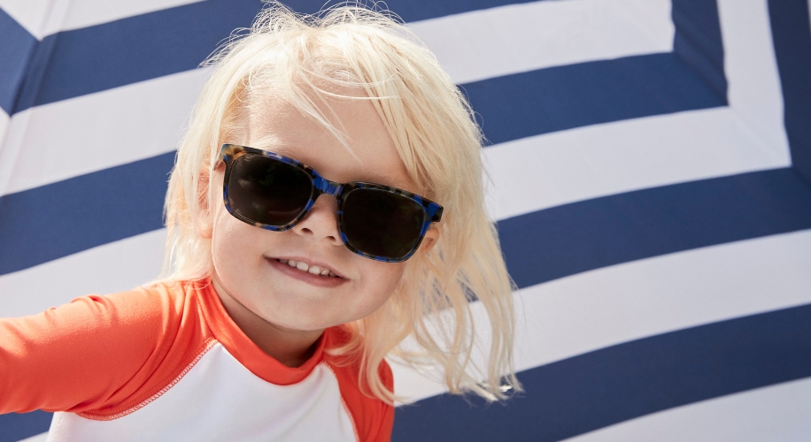 Child wearing tortoiseshell sunglasses and an orange-and-white rash guard shirt against a striped background.