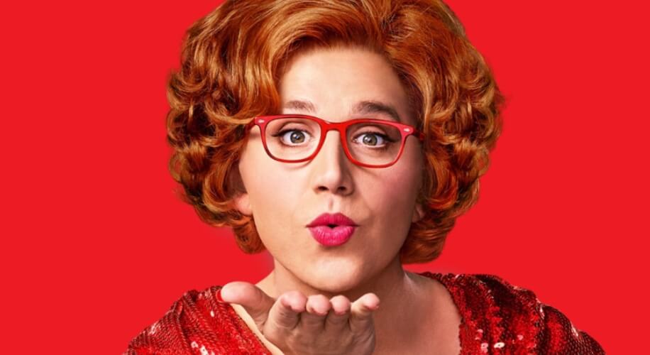 Person with short curly red hair and red glasses, wearing a red sequined top, blowing a kiss.